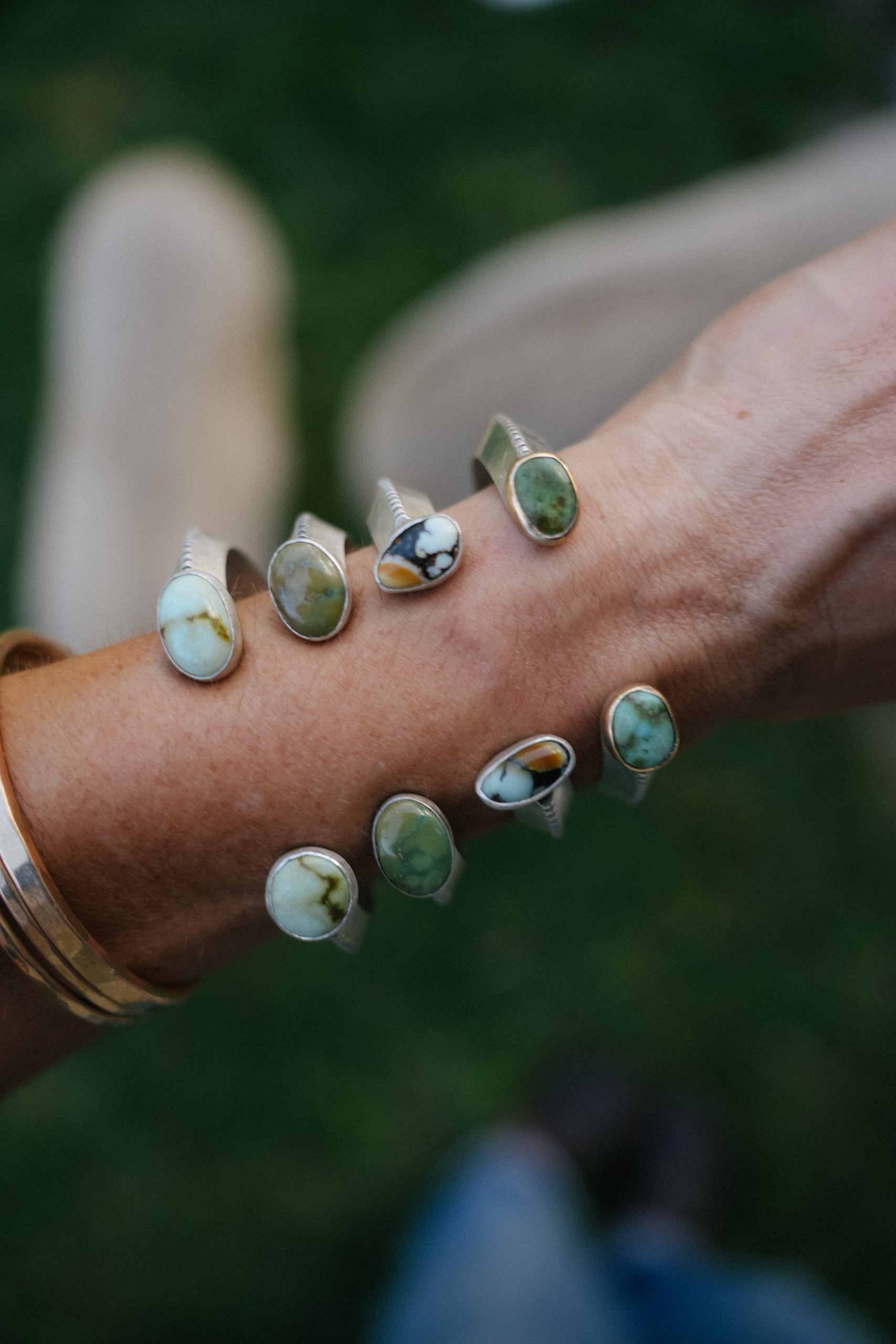 A close-up of a woman's wrist wearing multiple silver cuffs with oval variscite gemstones in green, white, and brown, set against a blurred outdoor background.