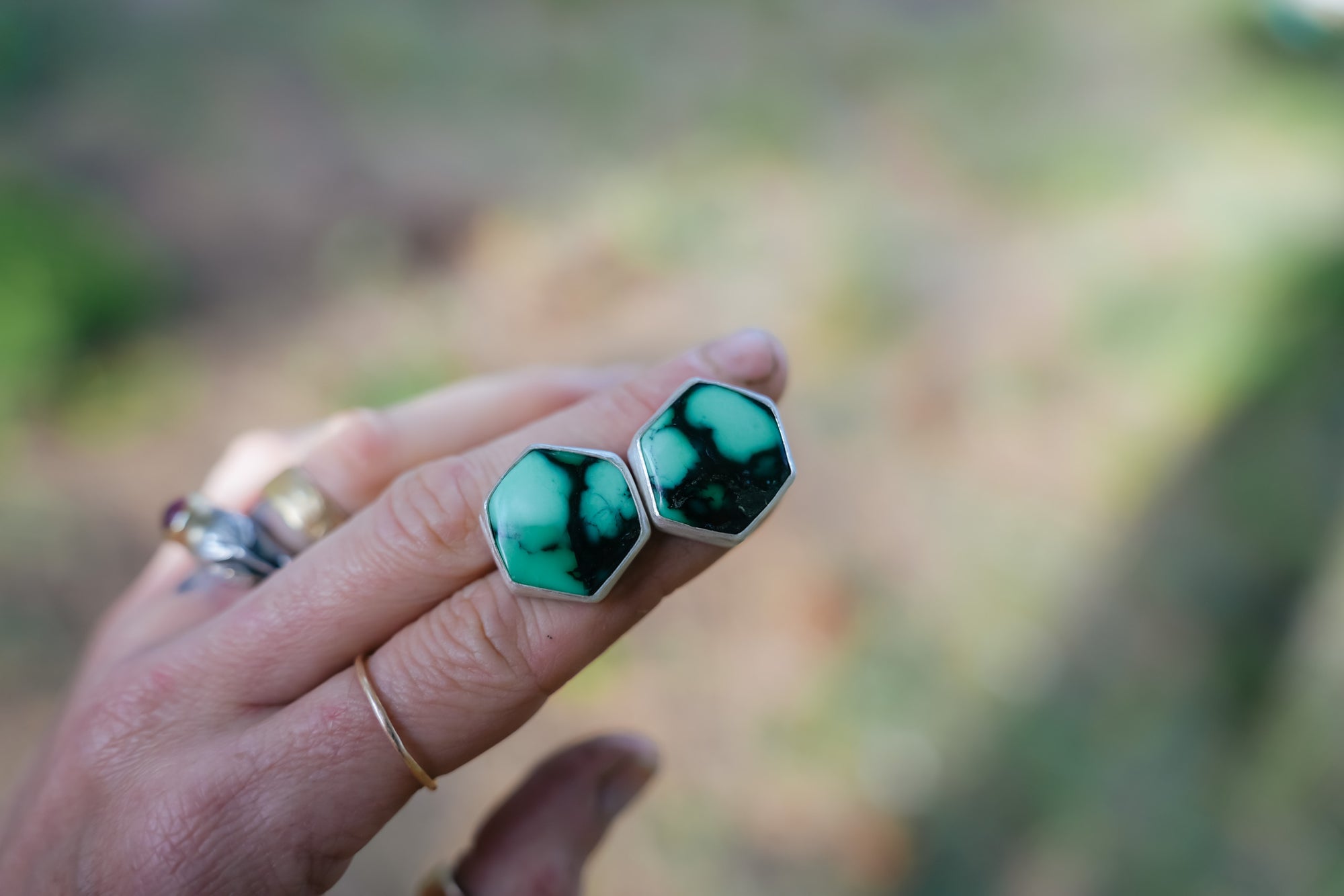 A woman's hand holding a pair of sterling silver and variscite post earrings. The gemstones are blue-green with black veining. The earring shape is hexagonal, and the natural background is blurred.