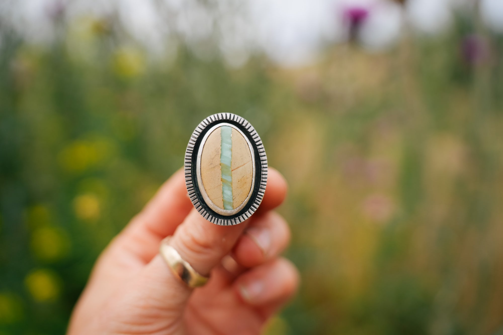 A woman's hand holding a sterling silver ring with a large, oval, variscite gemstone. The stone is cream-colored with a vertical band of turquoise color running through the center. The bezel is etched with vertical lines all the way around. The outdoor background of green foliage and yellow wildflowers. is blurred.