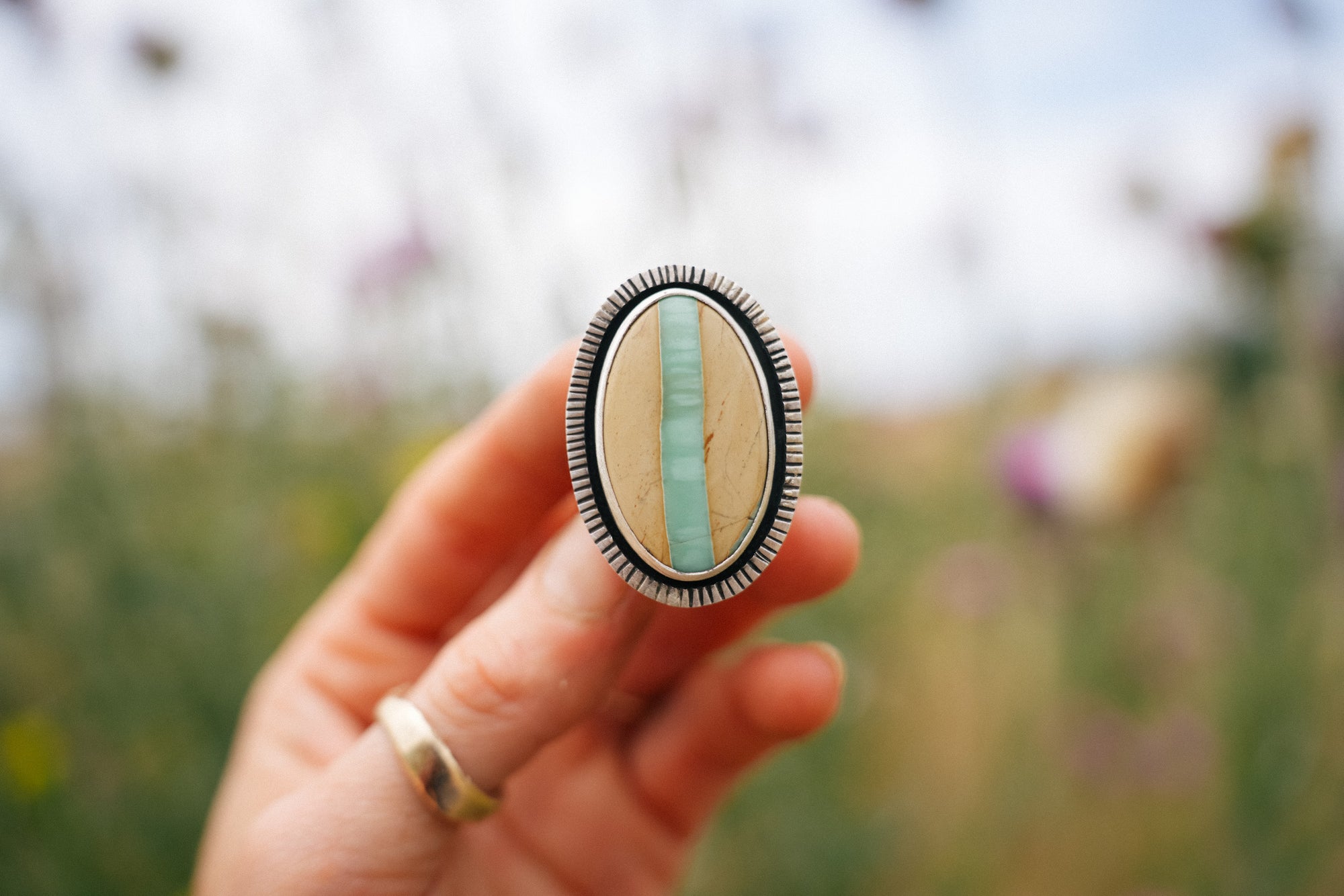 A woman's hand holding a sterling silver ring with a large, oval, variscite gemstone. The stone is cream-colored with a vertical band of turquoise color running through the center. The bezel is etched with vertical lines all the way around. The outdoor background of green foliage and lavender and purple wildflowers is blurred.