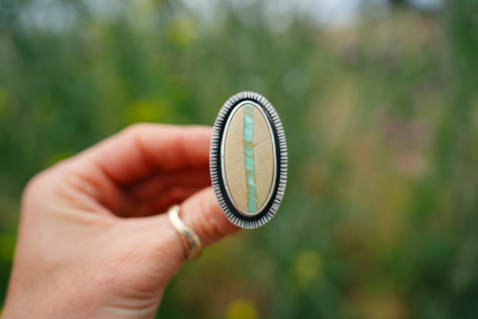 A woman's hand holding a sterling silver ring with a large, oval, variscite gemstone. The stone is cream-colored with a vertical band of turquoise color running through the center. The bezel is etched with vertical lines all the way around. The outdoor background of green foliage and yellow wildflowers. is blurred.