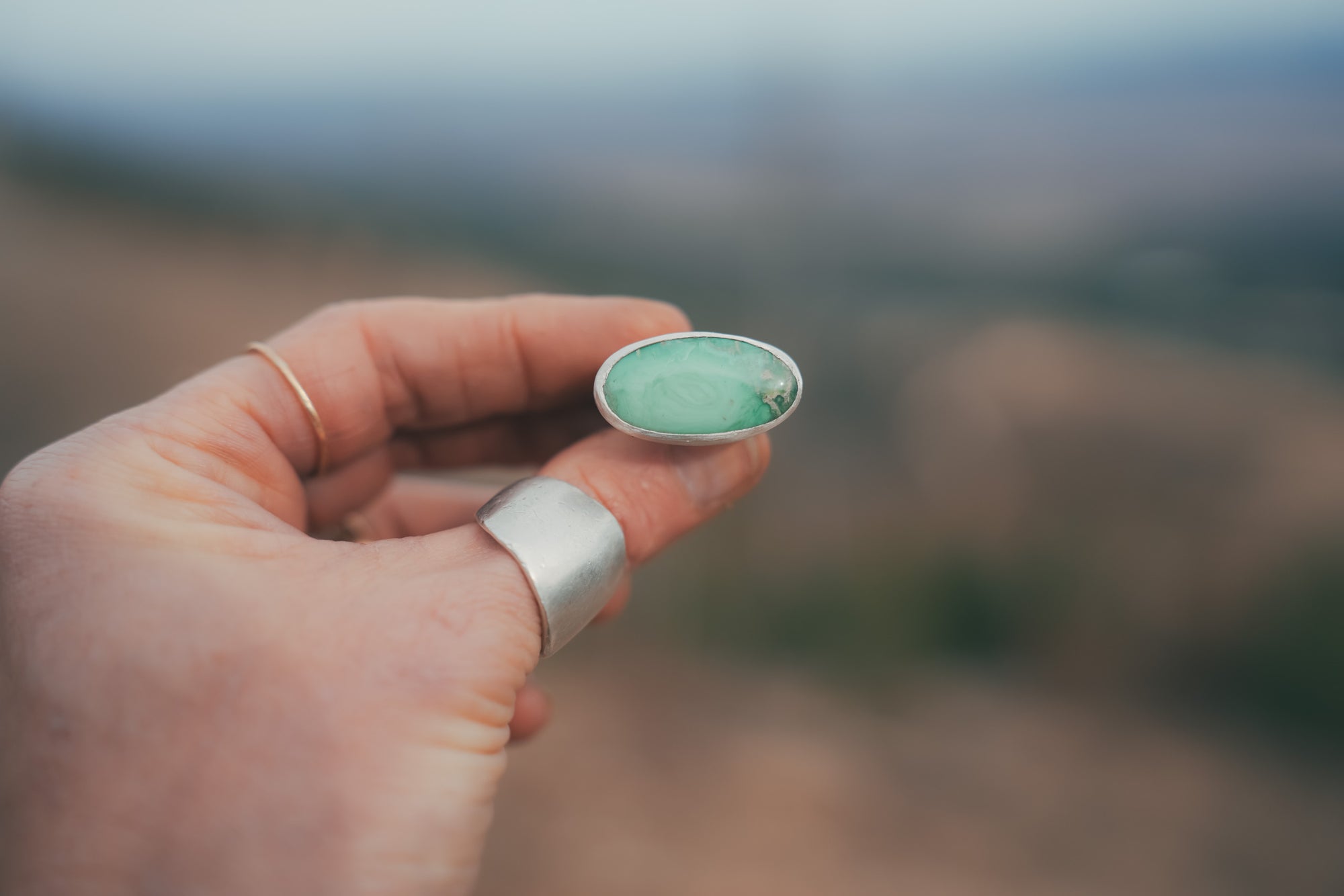 A woman's hand holding a sterling silver ring with an oval aqua-green gemstone against a blurred natural background.