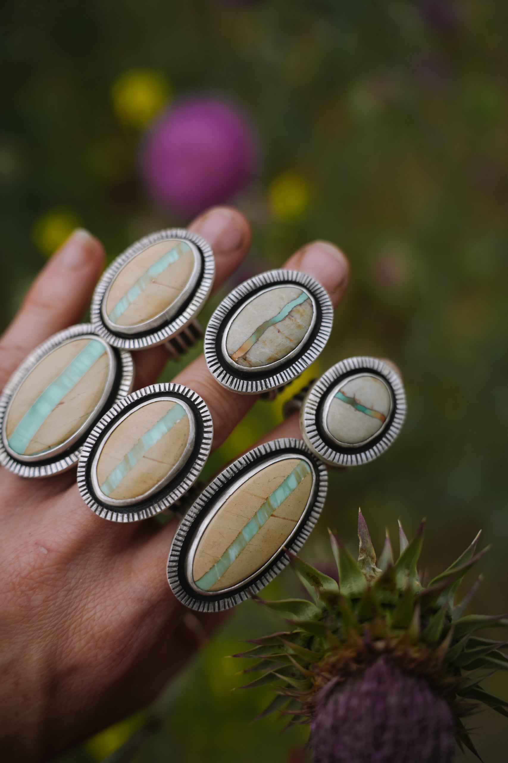 A woman's hand. She is wearing six large, bold sterling silver rings with oval turquoise and beige variscite gemstones. Photographed outdoors against a blurred green-and-purple natural background.