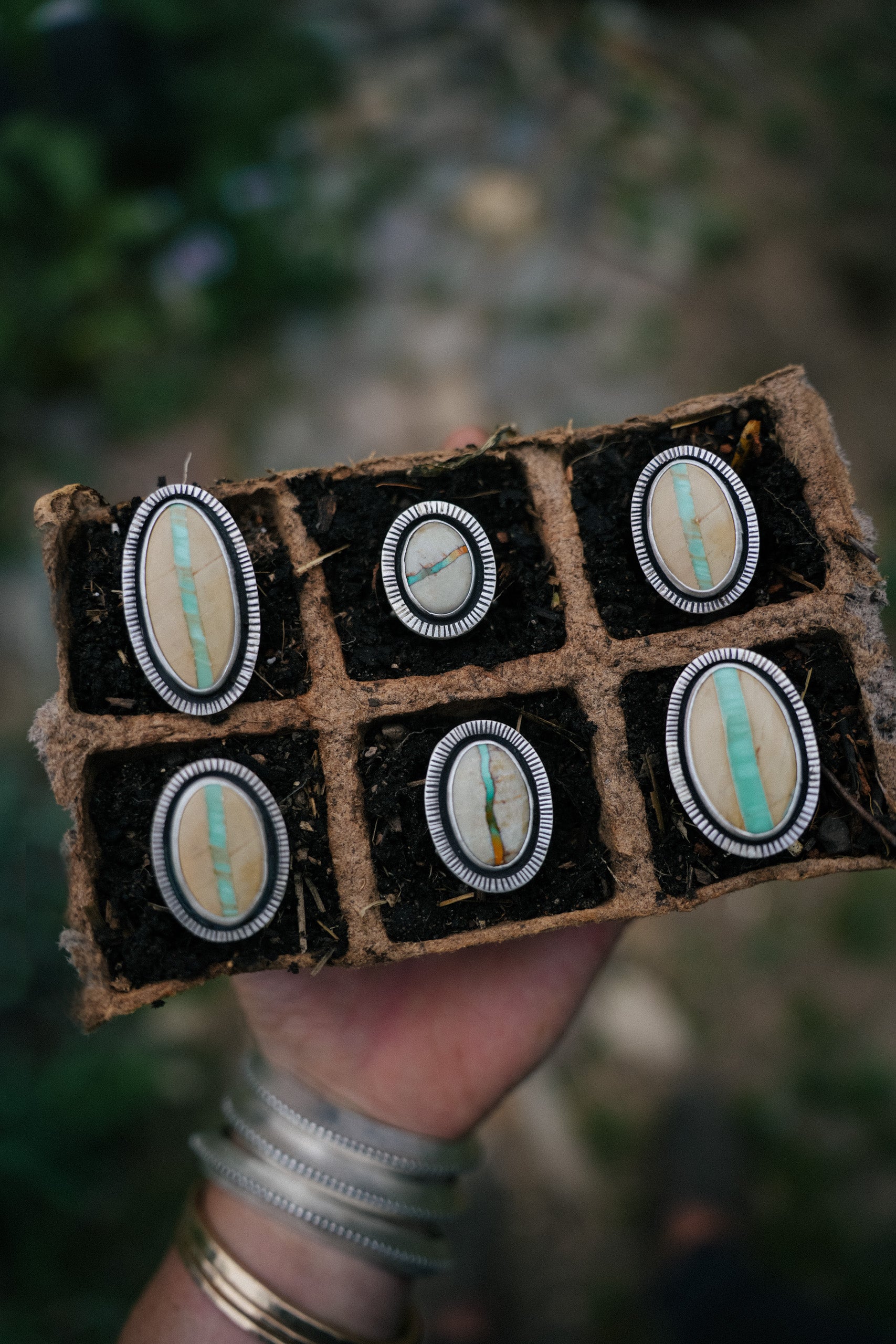 A woman's hand holding a small brown tray with six oval sterling silver and variscite rings set in a dark material.  Photographed outdoors against a blurred green and brown background. The woman is wearing silver bracelets.
