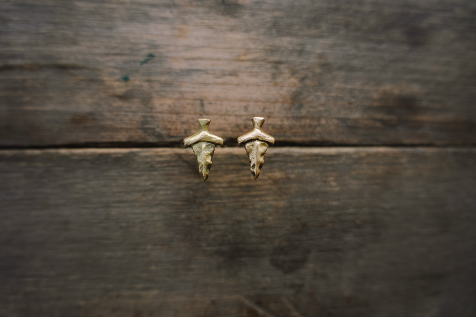 Small 14k gold, dagger-shaped post earrings against a blurred wooden background.