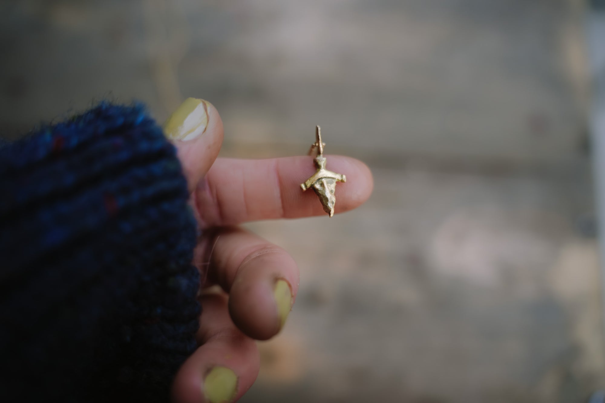 A small 14k gold, dagger-shaped pendant perched atop a woman's finger. She is wearing a dark blue sweater, and the natural background is blurred.