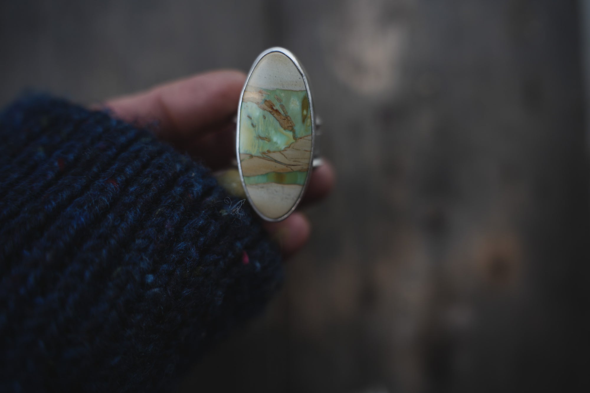 A woman's hand holding a sterling silver ring with an oval green-and-cream gemstone.