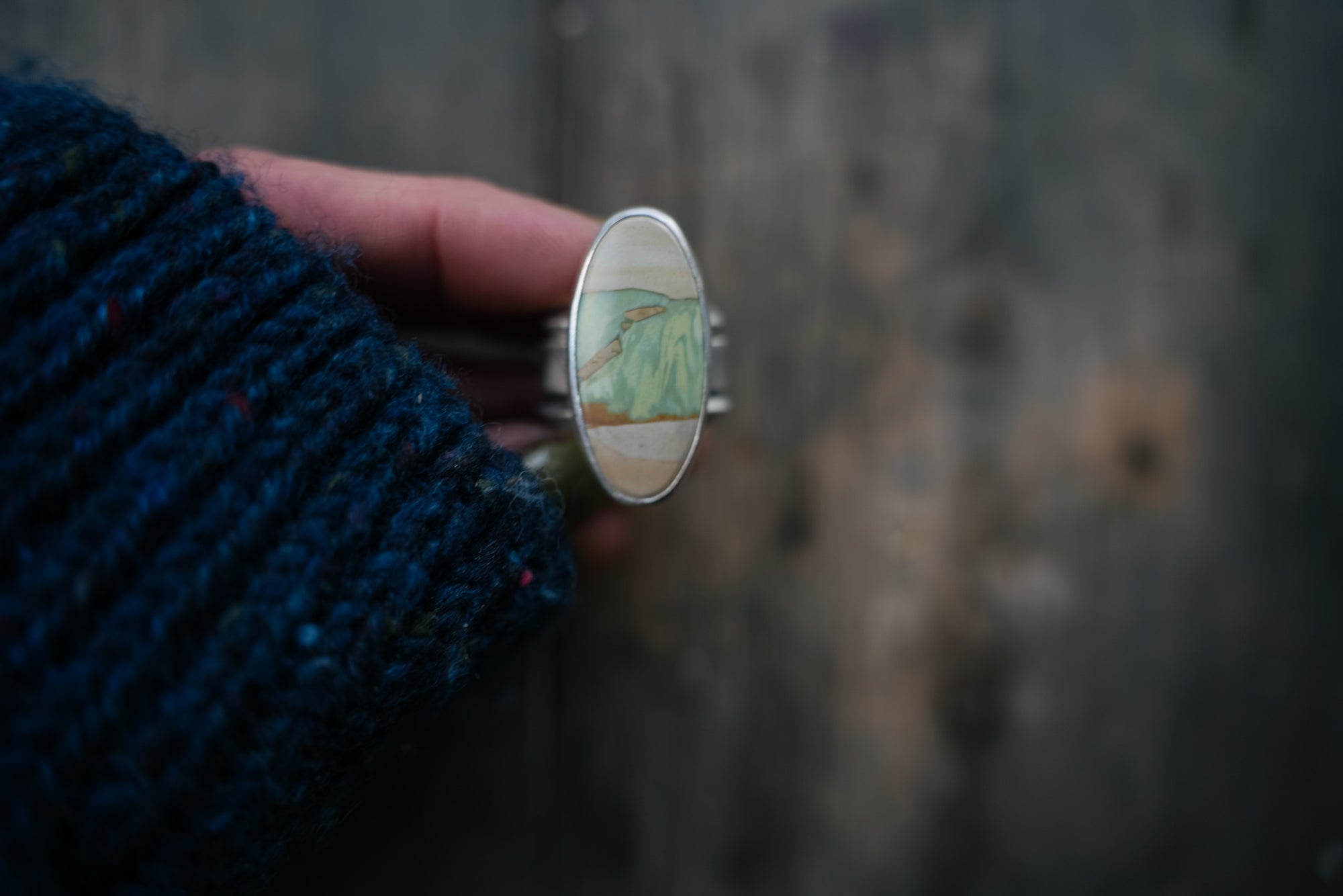 A woman's hand holding a sterling silver ring with an oval green, cream, and light brown variscite gemstone.