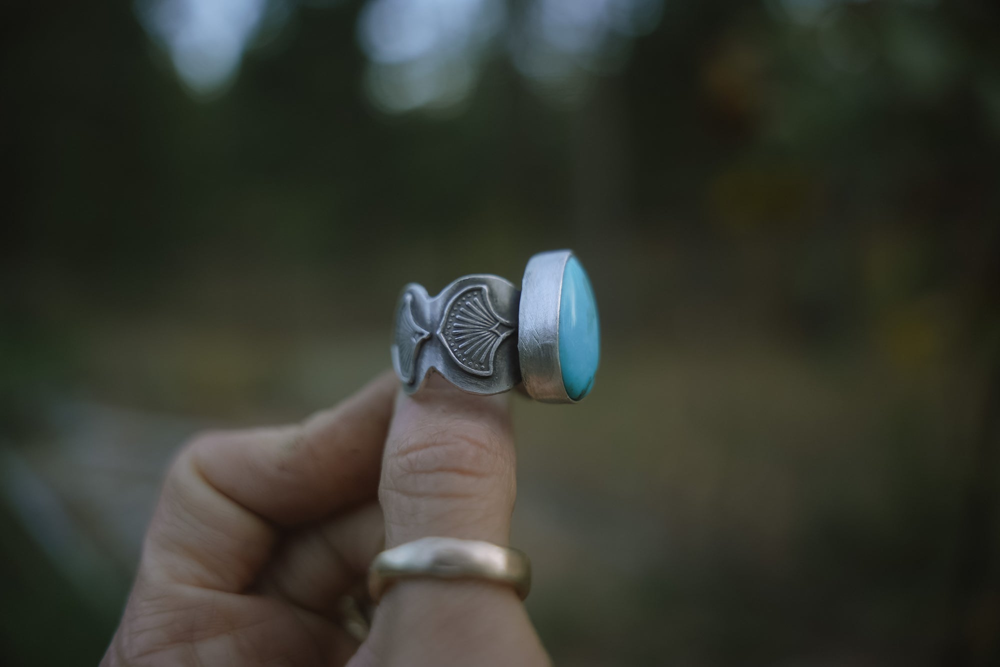 Sterling silver ring with a hand-stamped band and a turquoise stone held between fingers against a blurred natural background
