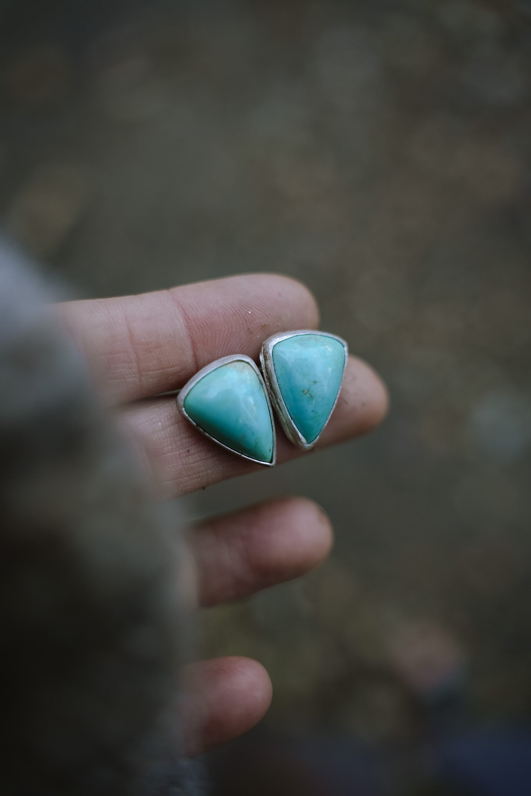 Triangular-shaped turquoise and sterling silver post-style earrings held in a woman's hand against a blurred background.