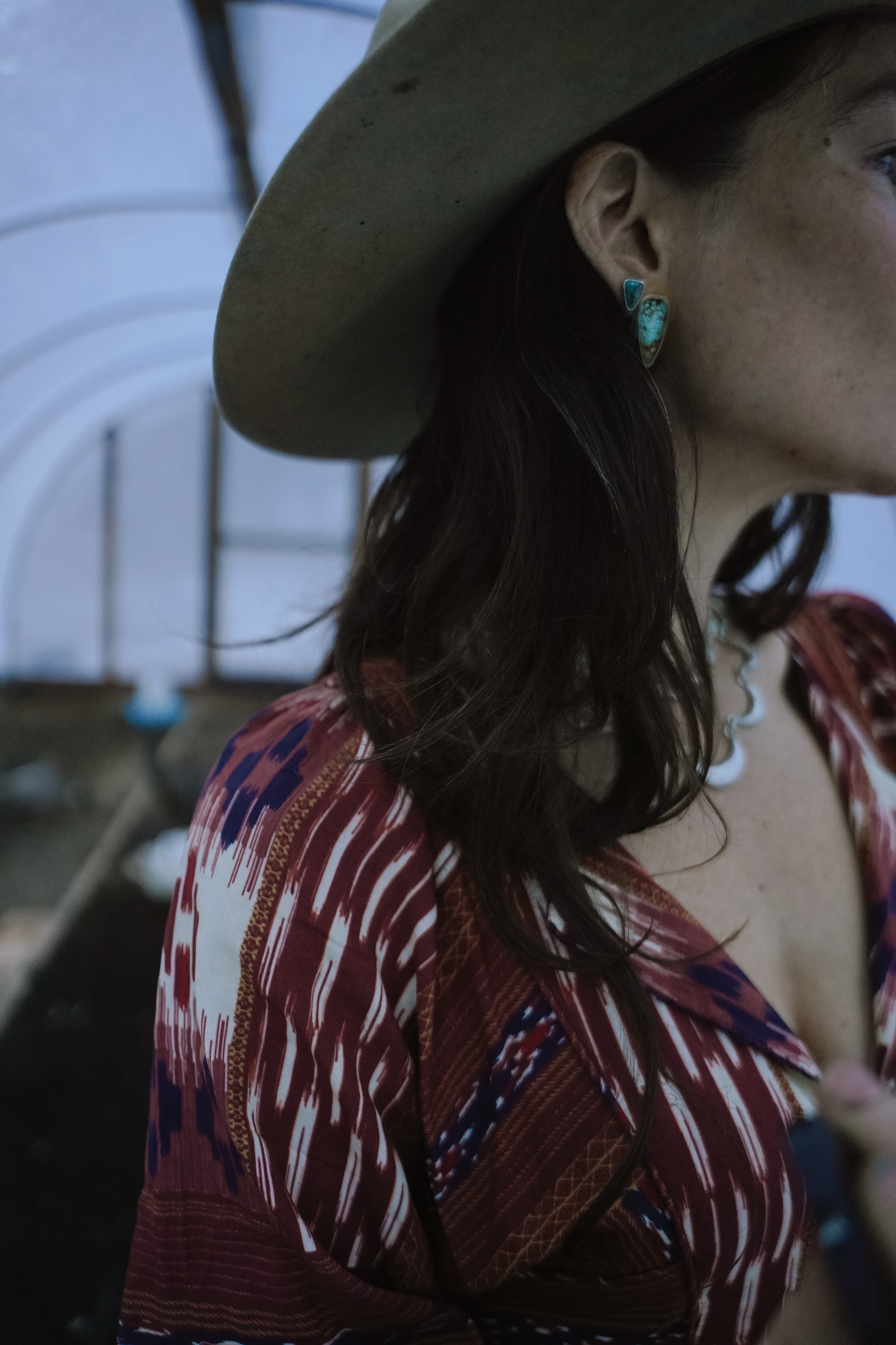 A woman with long dark hair wearing two turquoise spike-shaped earrings set in sterling silver against a blurred background. She is wearing a cowboy hat and a rusty-brown and white paterned dress.