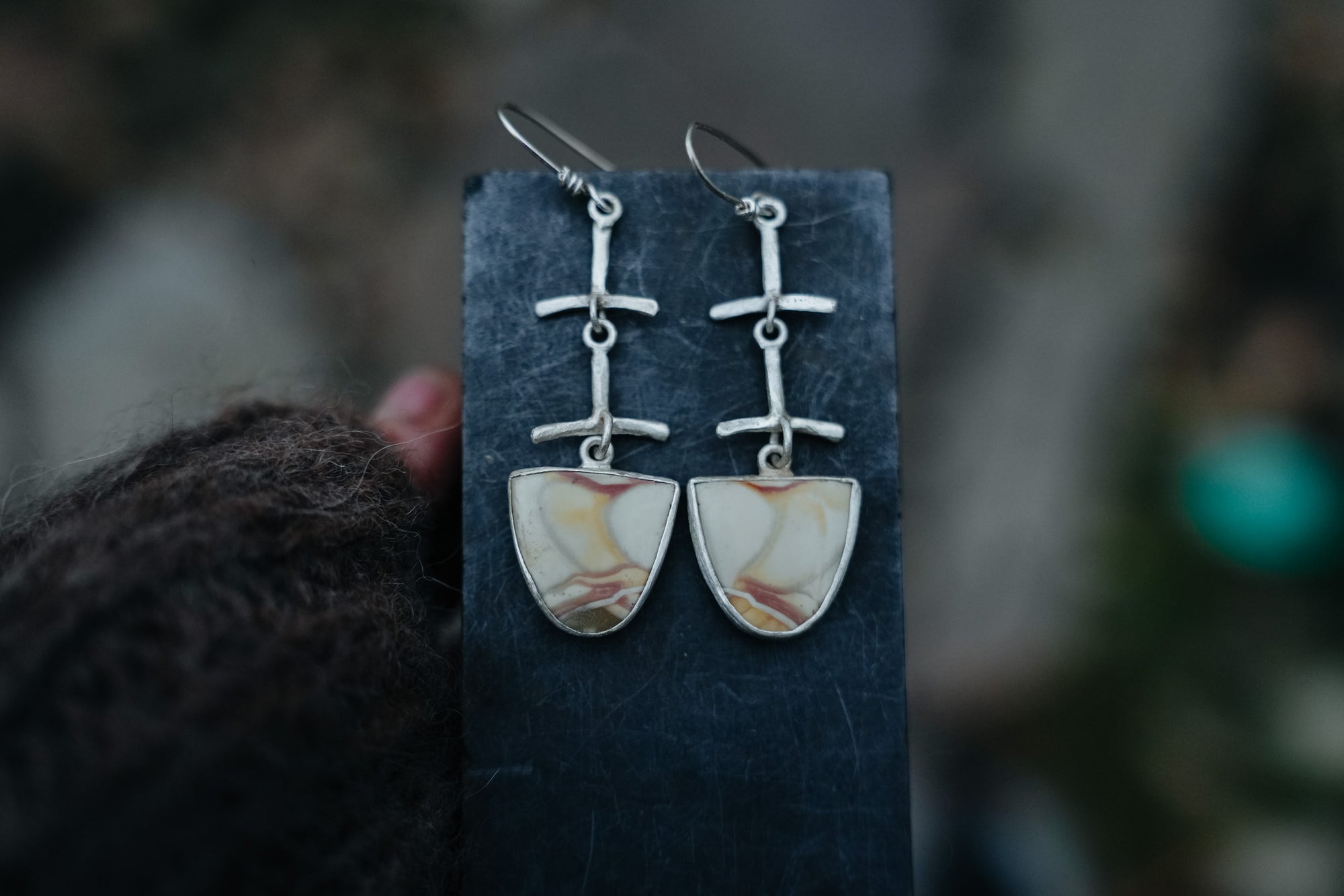 A pair of half-oval shaped sterling silver earrings with willow agate gemstones dangling from silver posts. They are hanging from a dark card held by a woman’s hand, against a blurred background.