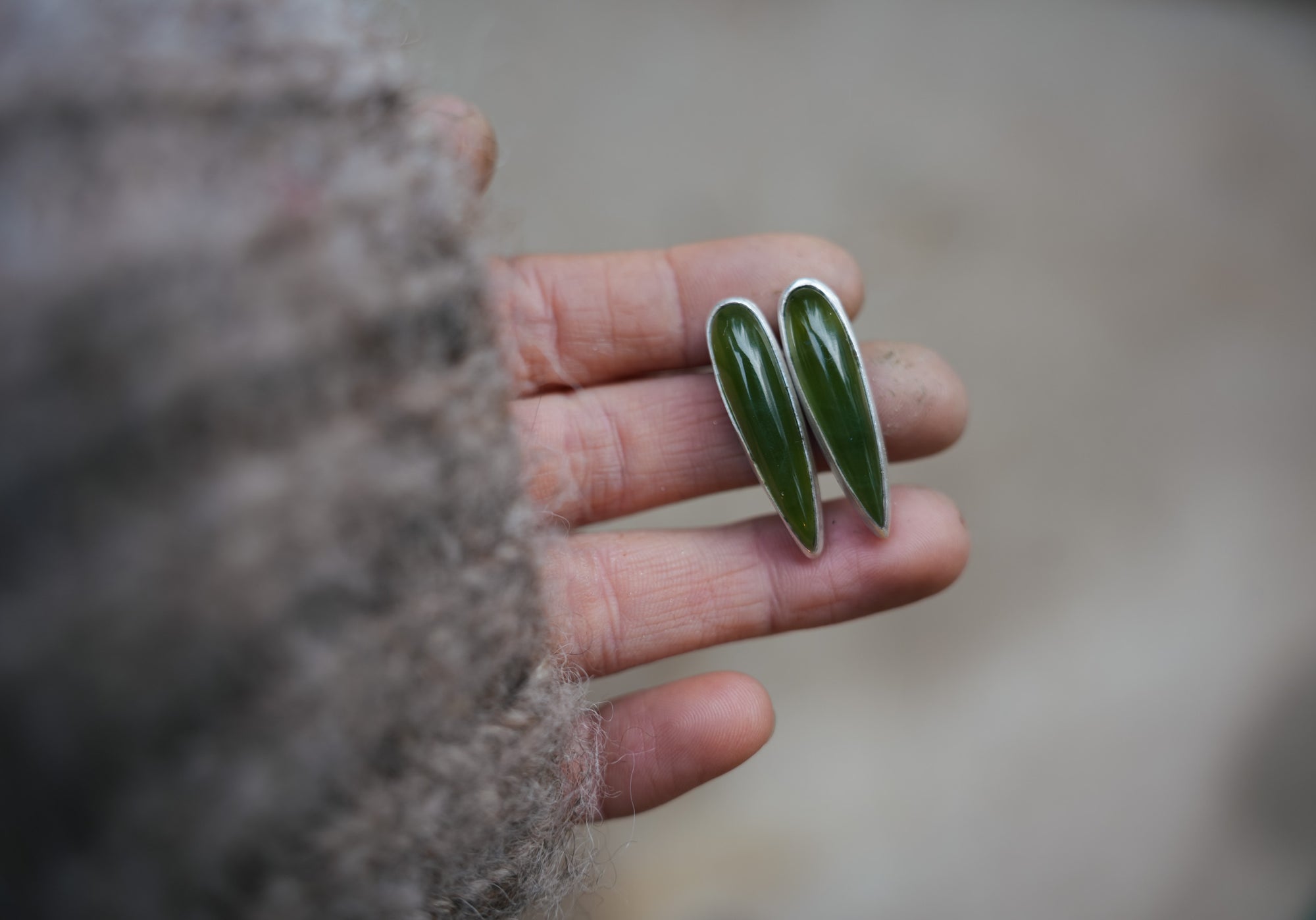 A woman's hand holding a pair of spike-shaped sterling silver and dark moss green vesuviante post earrings.