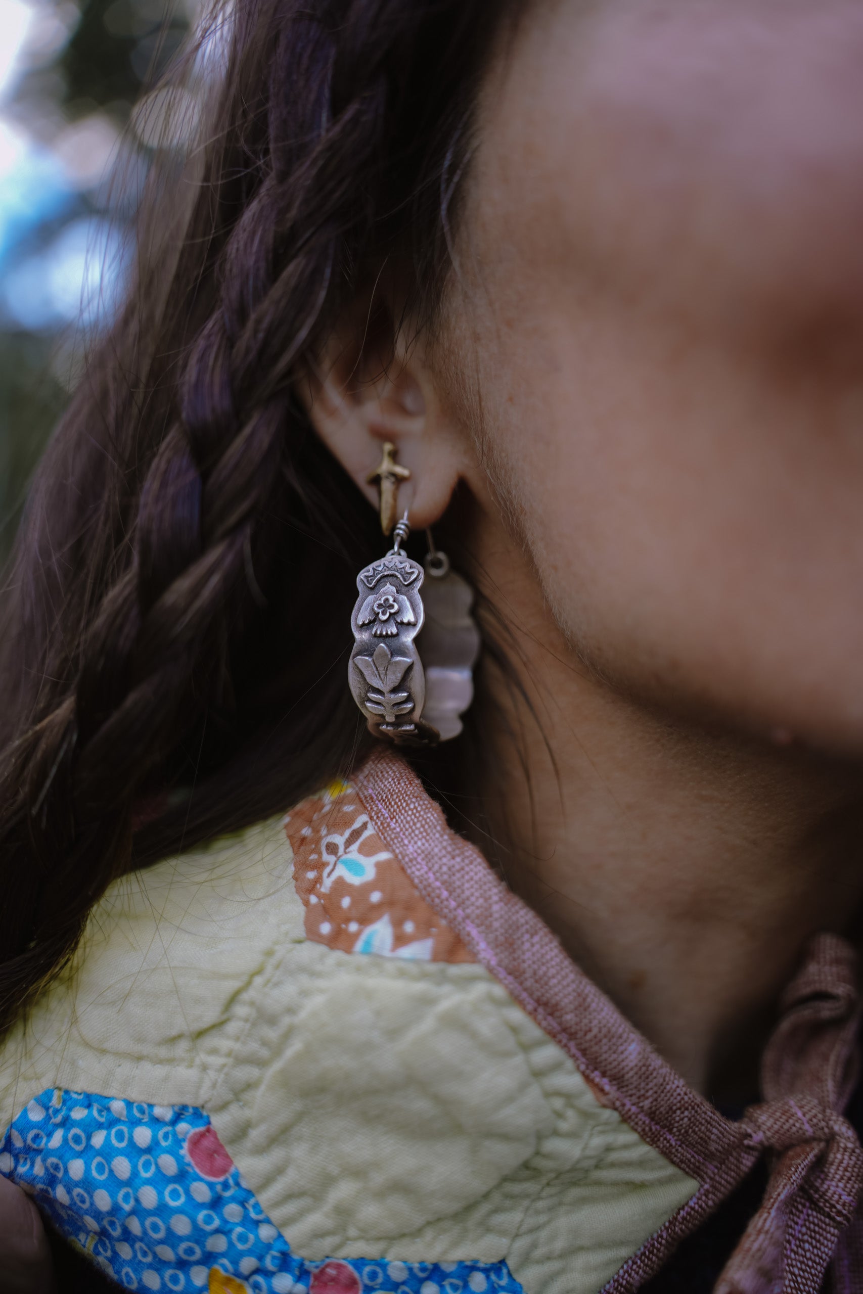 Close-up of a woman's ear with a large, wide-band sterling silver hoop earring. The earring body is adorned with raised, hand-carved icons including a flowering plant and a bird. The silver is oxidized creating a contrast, in areas, of black against silver. She has long, dark hair. 