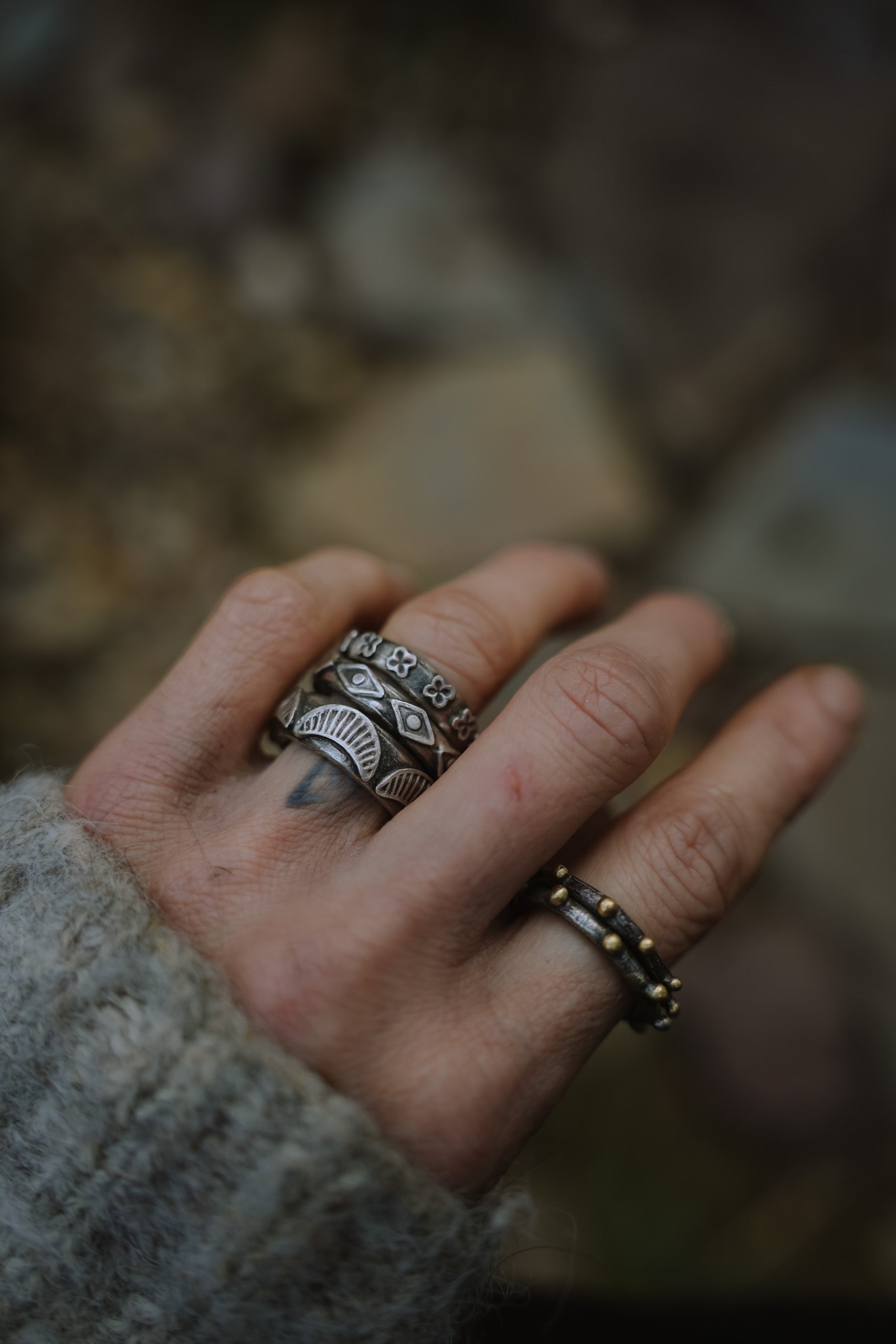 A woman's hand with a stack of wide-band sterling silver rings adorned with geometric shapes. She is also wearing two, thin oxidized sterling silver and 18K gold rings on her forefinger. The background is an outdoor setting and is slightly blurred.