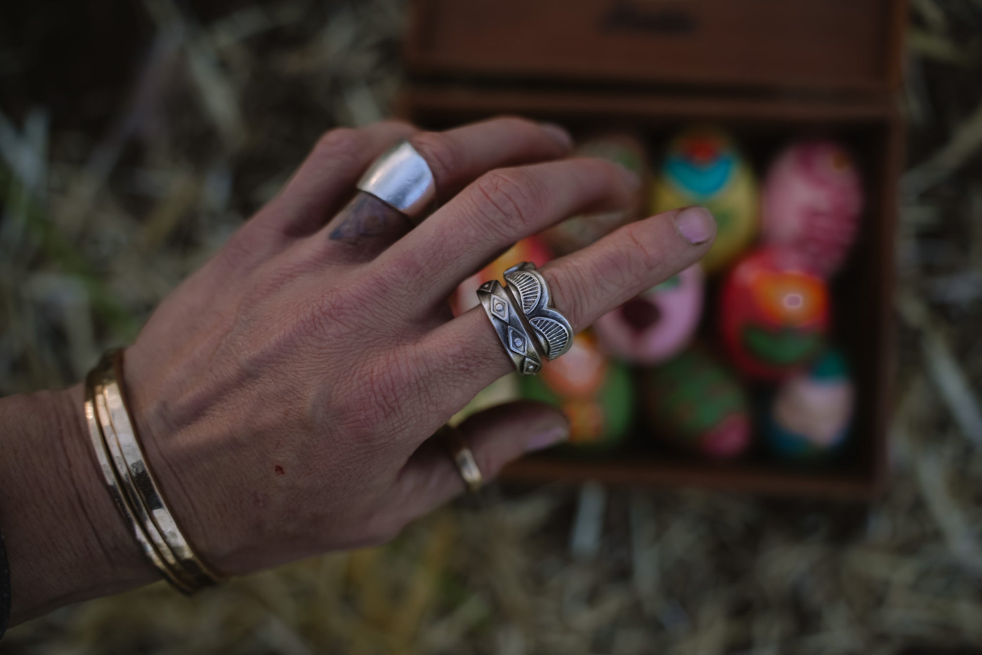 A woman's hand is wearing a stack of two wide-band sterling silver rings. The rings have shapes encircling the band. One with scallop shapes and the other with diamond shapes. The background is blurred, and a box of colorful objects lies beneath her hand.