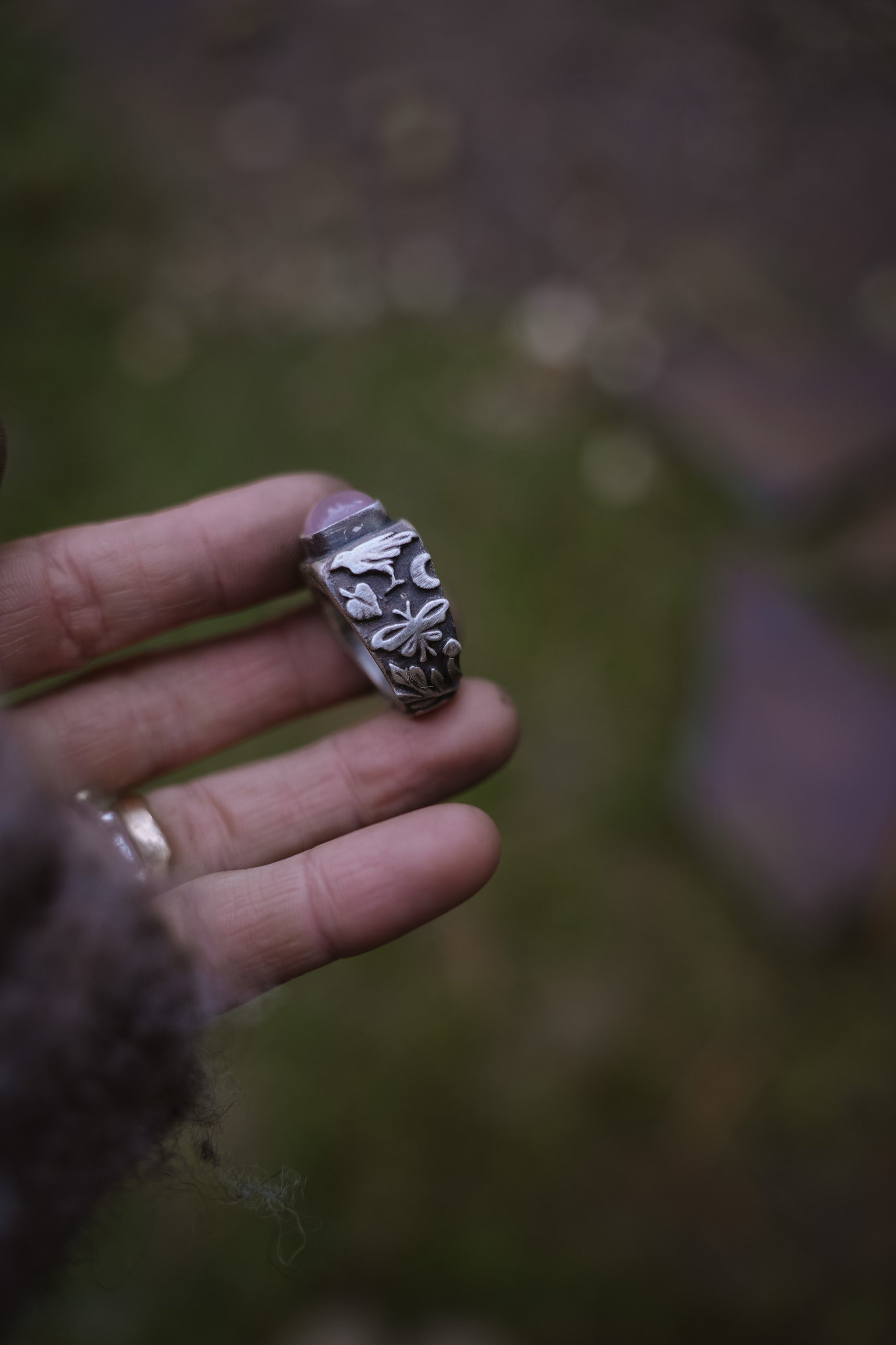A woman’s hand holding a sterling silver signet ring with a pink opal against a blurred natural background. Detail on the band shows a raven, a leaf, a butterfly, and a crescent moon.