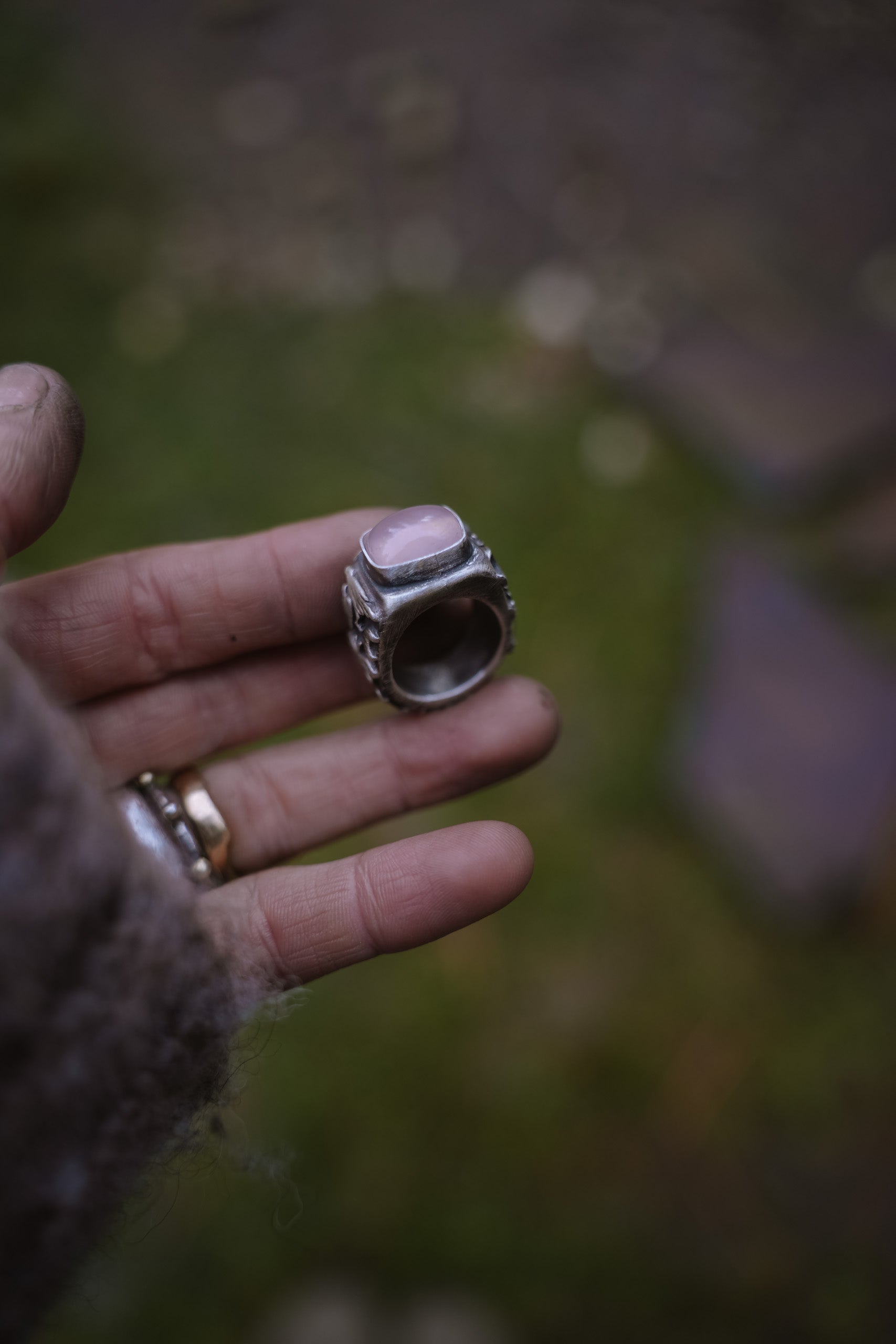 A woman’s hand holding a sterling silver signet ring with a pink opal against a blurred natural background.