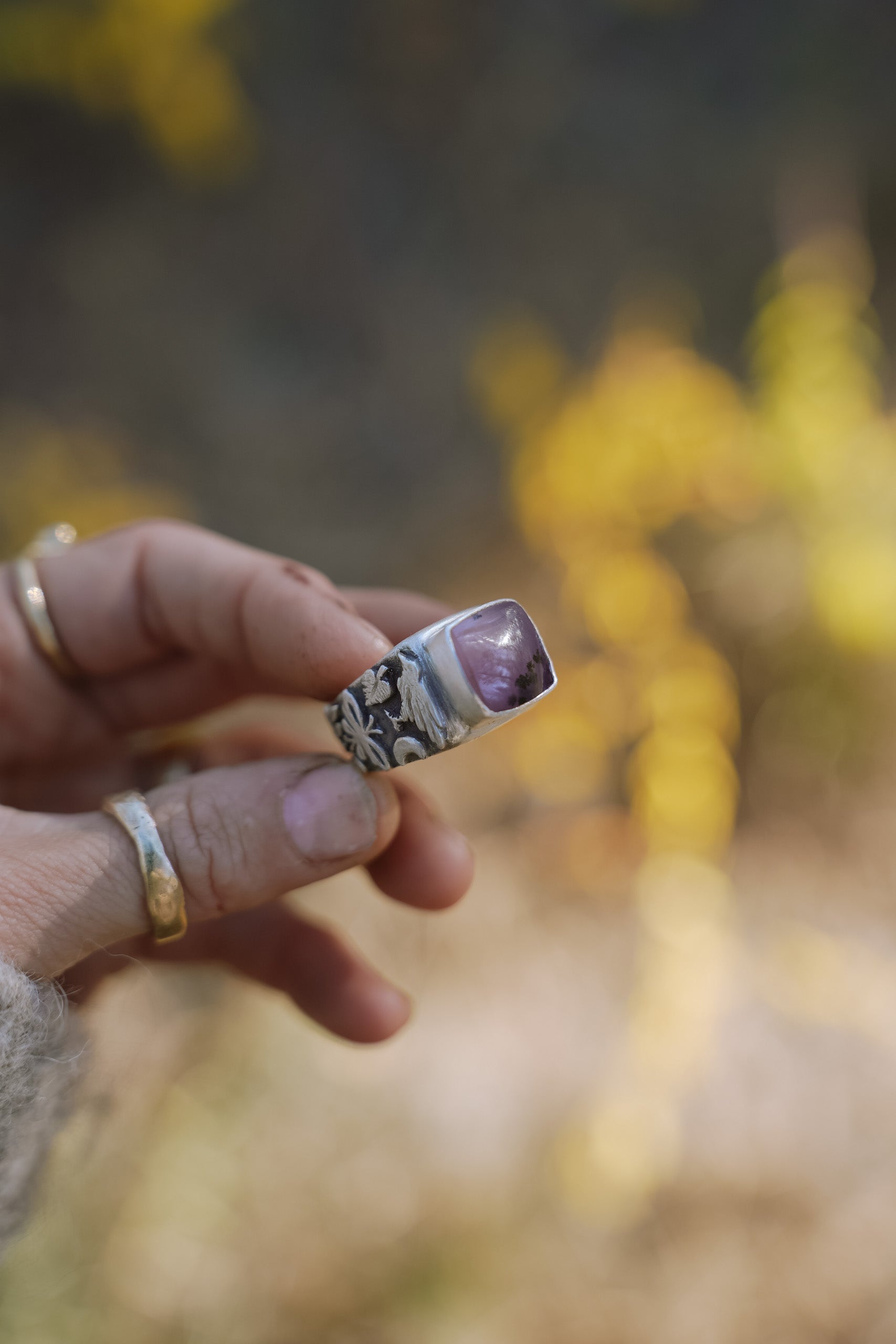 Woman’s hand holding a sterling silver signet ring with a pink opal against a blurred natural background. 