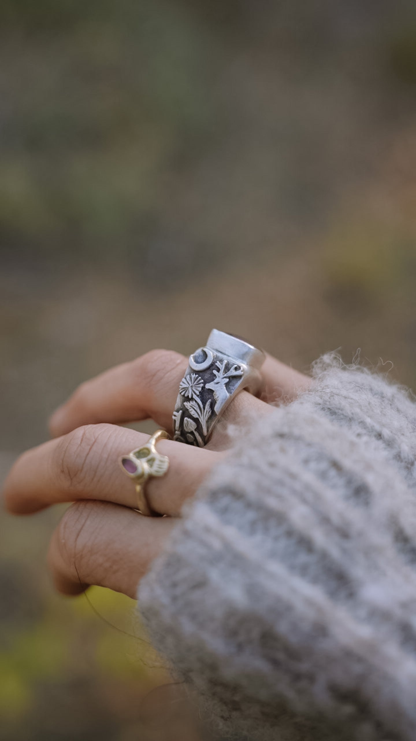 A woman’s hand wearing a sterling silver signet ring with a pink opal against a blurred natural background. Detail on the band shows a hand-carved moose, wildflowers, a mushroom, and a crescent moon.
