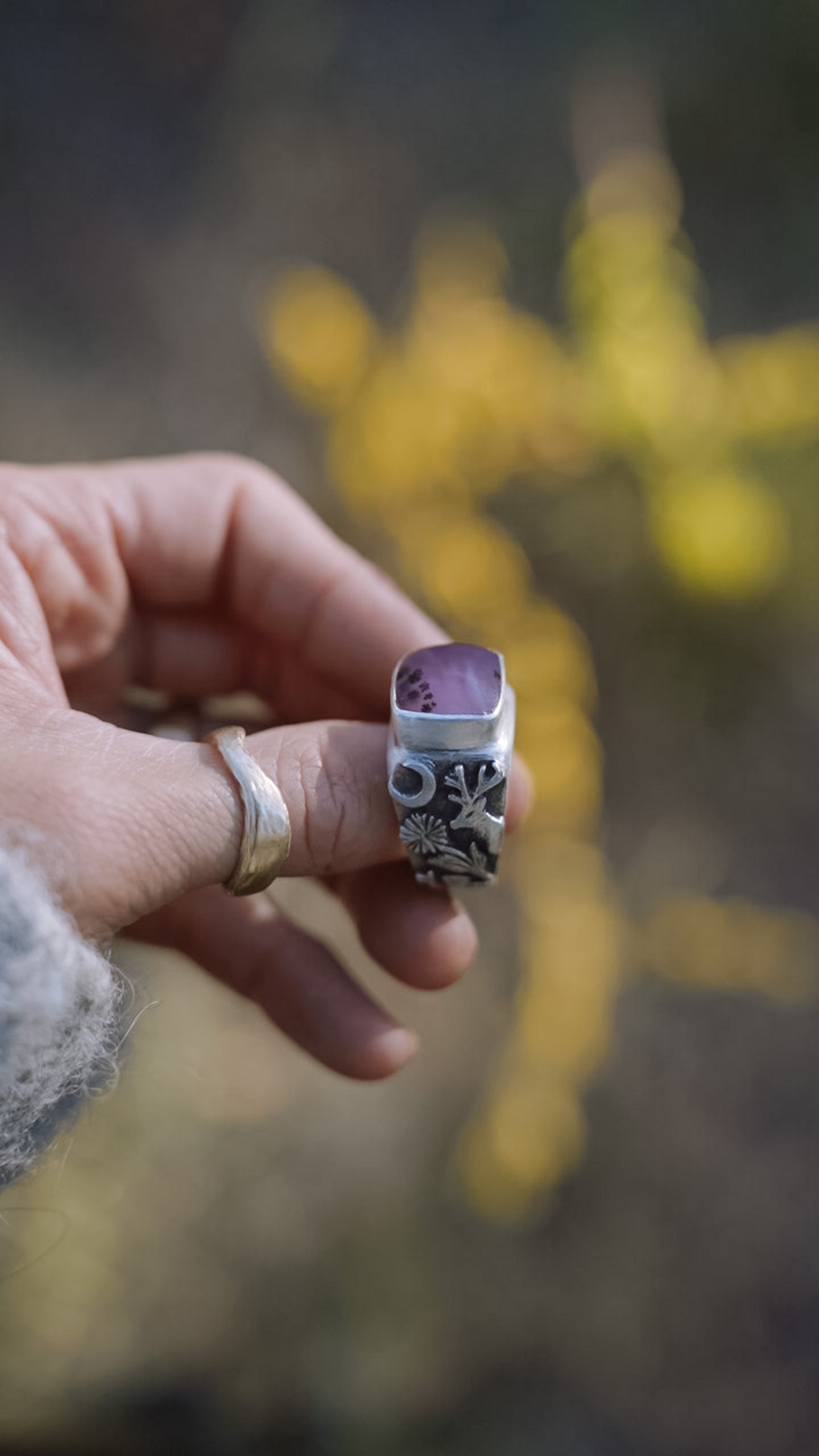 A woman’s hand holding a sterling silver signet ring with a pink opal against a blurred natural background. Detail on the band shows a hand-carved moose, wildflowers, a mushroom, and a crescent moon.