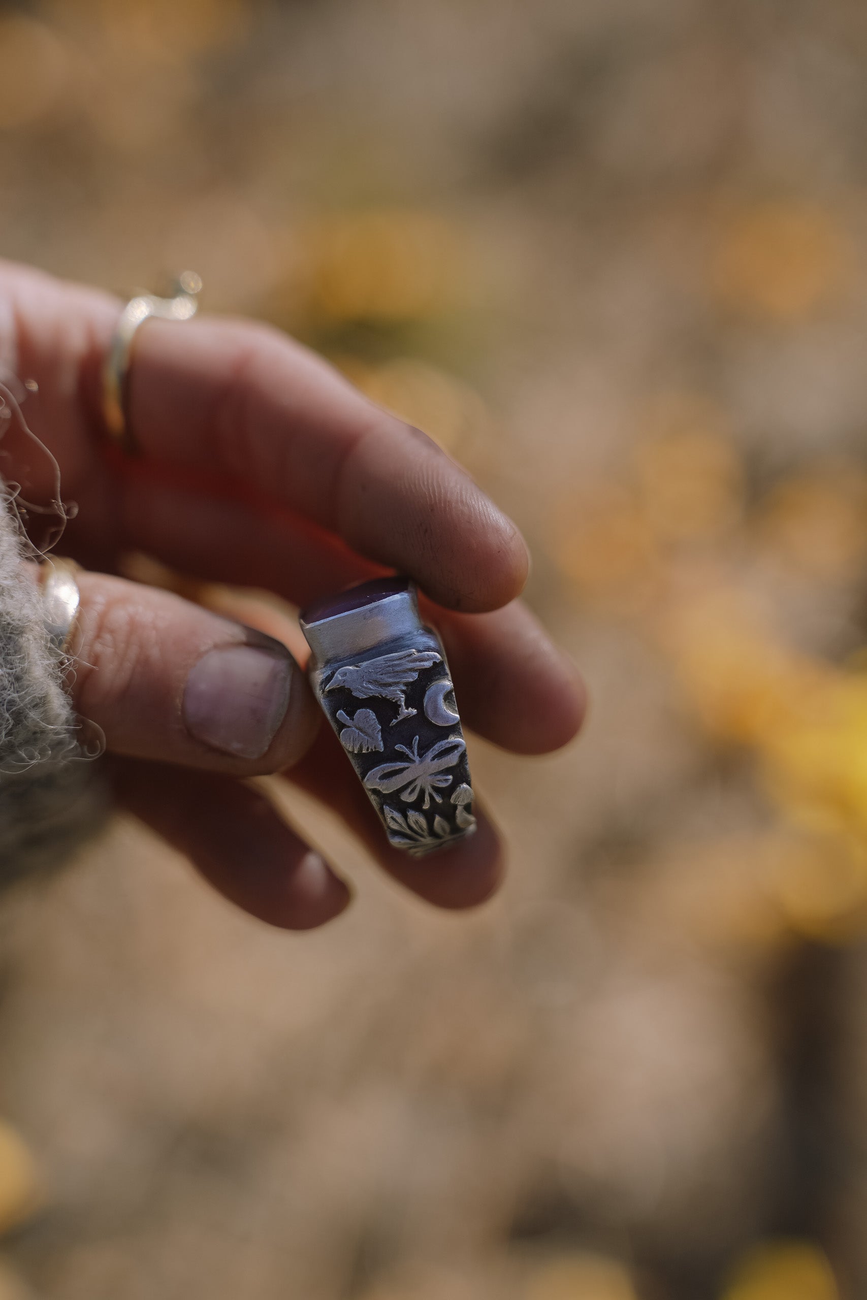 A woman’s hand holding a sterling silver signet ring with a pink opal against a blurred natural background. Detail on the band shows a raven, a leaf, a butterfly, and a crescent moon.