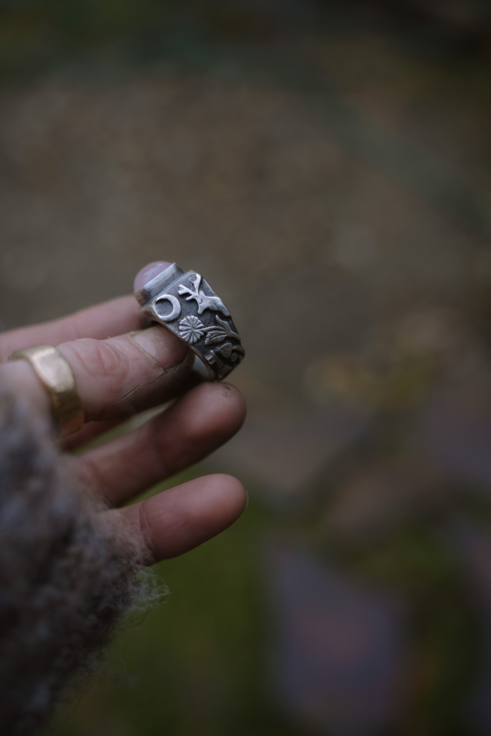 A woman’s hand holding a sterling silver signet ring with a pink opal against a blurred natural background. Detail on the band shows a hand-carved moose, wildflowers, a mushroom, and a crescent moon.